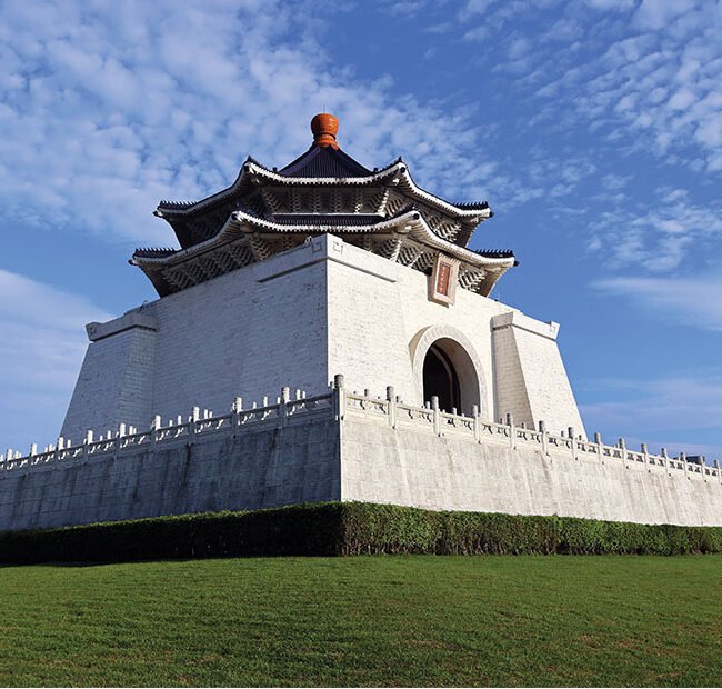 White stone Chinese-style gate on a raised platform with a curved tiled roof, arch entrance, white balustrade, and a green lawn under a blue sky (watermark: GB Giving Back)
